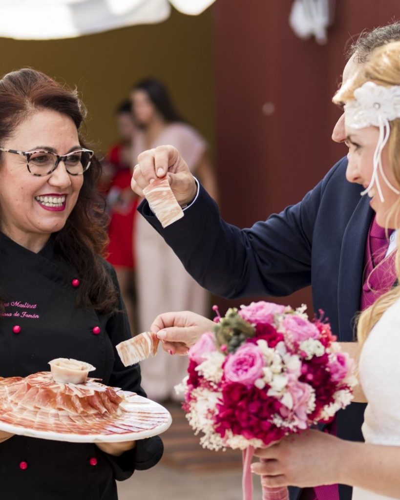 Cortadora de jamón risueña y amable ofreciendo un plato de jamón con forma de rosa a dos novios en una boda.